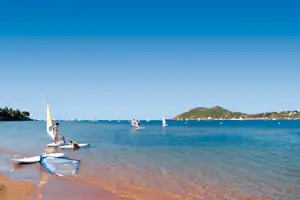 Windsurf-Boards am Strand, Segelboote auf dem blauen Wasser und Küste mit Hügeln im Hintergrund.