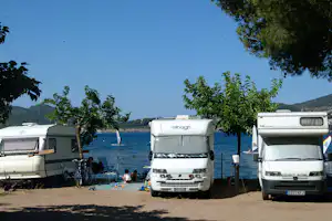 Mehrere Wohnmobile auf einem Campingplatz am See, mit Bäumen im Vordergrund und Stellplätzen am Ufer.