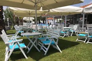Weiße Campingstühle mit blauen Auflagen unter Sonnenschirmen auf dem Rasen, im Hintergrund weitere Sitzplätze und Gebäude.