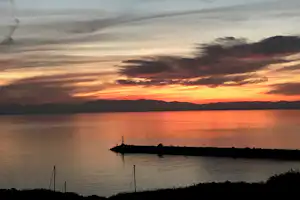 Sonnenuntergang über einem See mit spiegelndem Wasser, dunkler Bergkette am Horizont und bewölktem Himmel.