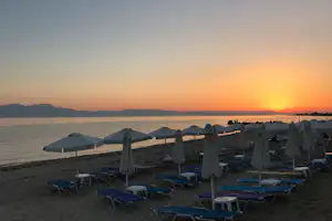 Reihe blauer Liegen mit Sonnenschirmen am Strand, dahinter das Meer und ein Sonnenuntergang am Horizont.