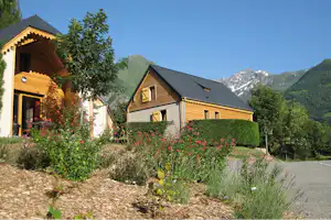 Zwei Holz-Chalets auf dem Campingplatz mit Blumenbeet, Hecke und Blick auf die Berge im Hintergrund.