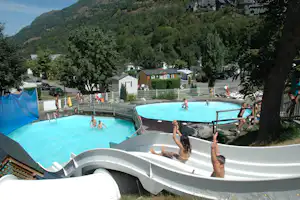 Campingpool mit Schwimmern, dahinter Bungalows und Wohnwagen, umgeben von Bäumen und einem Berg im Hintergrund.
