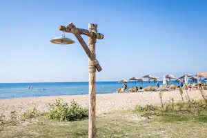 Strand mit hölzernem Pfahl, Sonnenschirmen und Liegen am Meer, im Hintergrund blaues Wasser und Himmel.
