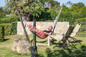 Mann liegt in einer bunten Hängematte auf dem Campingplatz, daneben Liegestuhl und Steinmauer im Grünen.