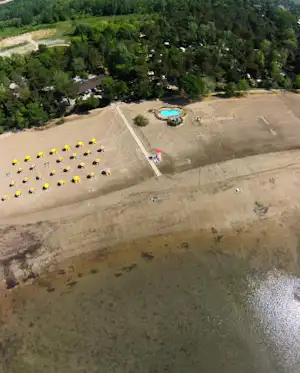 Luftaufnahme eines Campingplatzes am See mit Pool, Stellplätzen, Wegen und Bäumen im grünen Umland.