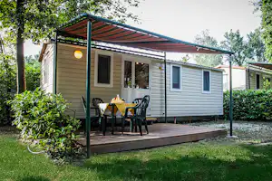 Wohnwagen mit überdachter Terrasse, Tisch und Stühlen, umgeben von Hecken und Bäumen im Campingplatz-Grün.