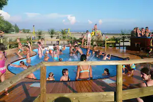 Kinder und Erwachsene planschen im Pool auf einer Holzterrasse, mit Blick auf das Meer und Palmen im Hintergrund.