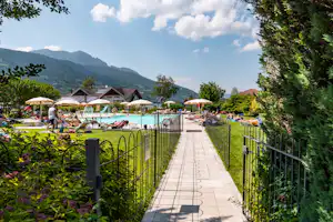 Gepflegter Weg am Campingplatz entlang eines Pools, mit Zäunen, Liegen und Blick auf Berge und Bäume.