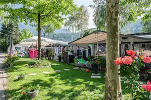 Campingplatz mit Rasenfläche, Zelten und Wohnwagen-Stellplätzen, Tischen und Stühlen unter Sonnenschirmen zwischen Bäumen.