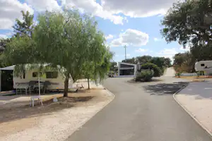 Campingplatzweg mit Kies, Bäumen und Wohnwagen, daneben ein Gebäude und weitere Stellplätze.