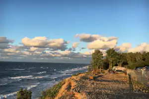 Küsten-Campingplatz am Meer mit Grasweg, Bäumen, Wohnwagen und Autos am Zaun, im Hintergrund Wellen und Wolken.