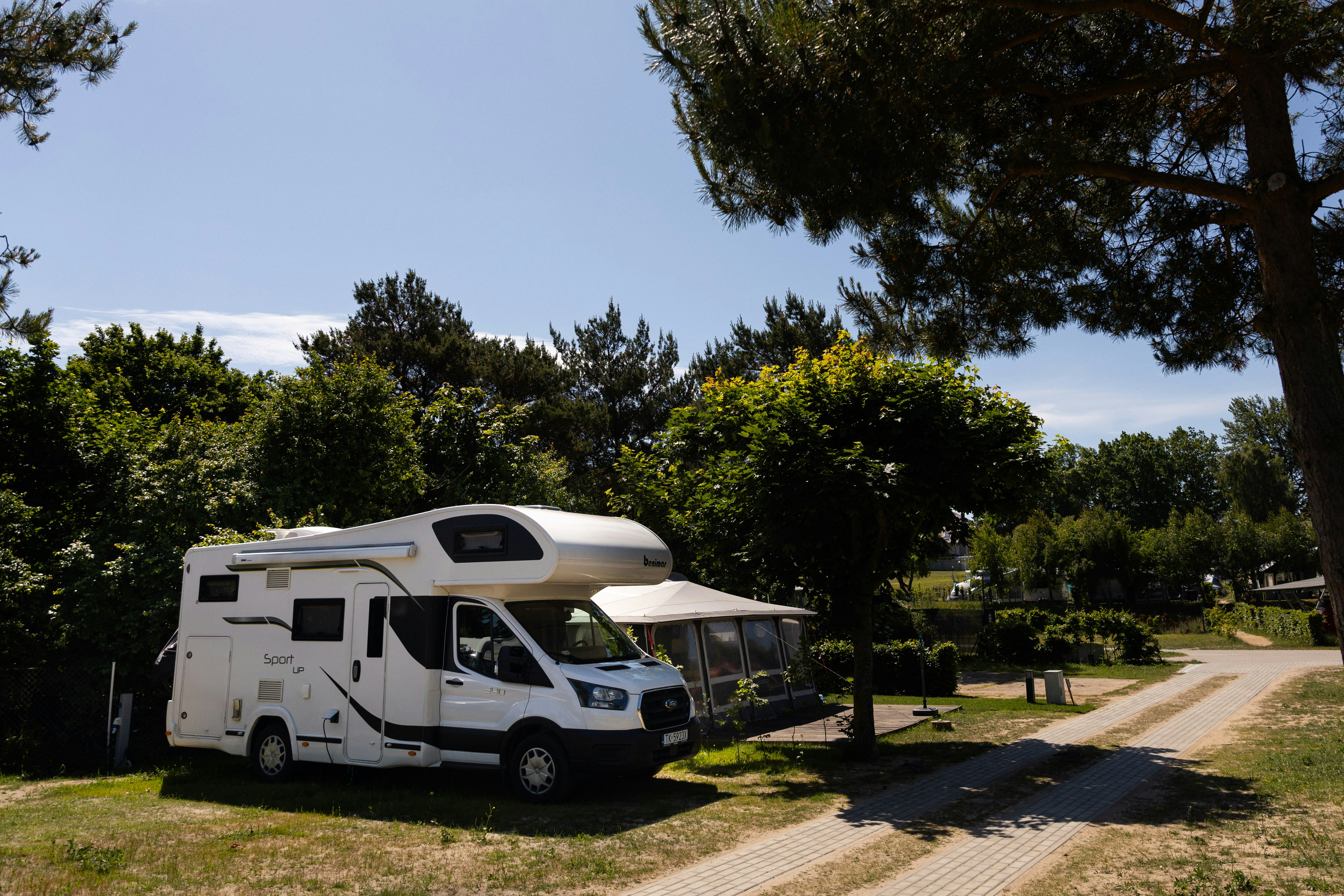 Wohnmobil auf einem Campingplatz am Weg, neben Bäumen und Hecken, mit Rasenfläche und Stellplatzmarkierung.
