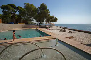 Schwimmbecken am Campingplatz mit Blick auf das Meer, Sandstrand, Palmen und Liegen am Ufer.
