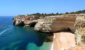 Steilküste mit Felsbögen und türkisfarbenem Wasser, darunter ein kleiner Sandstrand am Meer.