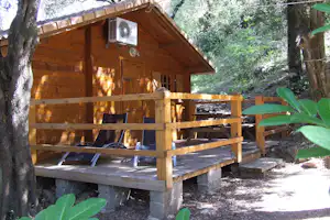 Holzhütte mit Terrasse, Holzgeländer und zwei Stühlen vor dem Eingang, umgeben von Bäumen im Campingbereich.