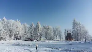 Verschneite Campinganlage mit zugefrorenem See, Holzsteg und kleinen Gebäuden im Hintergrund, umgeben von verschneiten Bäumen.