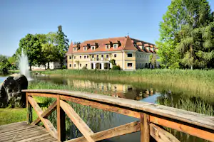 Hotelähnliches Gebäude am See mit Springbrunnen, Schilf und Holzgeländer im Vordergrund, umgeben von Bäumen.