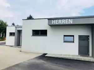 Gebäude mit Beschriftung „HERRREN“, Eingangstür und Fensterfront, gepflasterter Weg und Betonrampe am Campingplatzgelände.