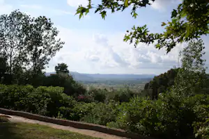 Blick vom Campingplatz ins Tal mit Feldern, Hügeln und Bäumen im Vordergrund, umrahmt von Grün und Blumen.