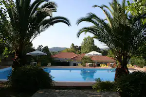 Pool am Campingplatz mit Palmen, Liegestühlen und Sonnenschirmen, dahinter Gebäude mit roten Ziegeldächern und Garten.