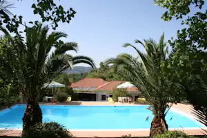 Blauer Pool mit Liegestühlen, Palmen und Blick auf ein Haus mit rotem Ziegeldach und Sonnenschirmen.