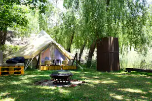 Zelt auf einem Campingplatz im Grünen unter Bäumen, mit Grill und Sitzbank im Vordergrund.