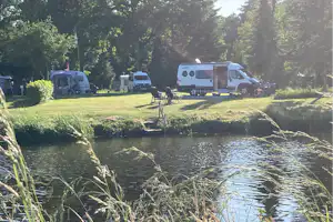 Campingplatz am Fluss mit Rasenfläche, Caravanen und Wohnwagen, Sitzgruppe am Ufer und Bäumen im Hintergrund.