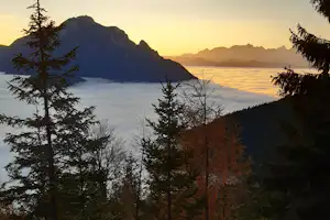 Blick auf Berge und einen See bei Sonnenaufgang, umgeben von Tannen und bewaldeten Hängen.