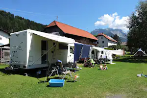 Wohnwagen auf einem Campingplatz auf grünem Rasen, mit Campingstühlen, Tisch und Wäscheleine vor Häusern und Bergen.