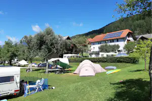 Campingwiese mit mehreren Zelten, Wohnwagen und Campingstühlen, im Hintergrund Häuser mit roten Dächern und Bäume.