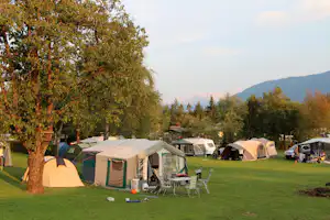 Zelte auf einem grünen Campingplatz, daneben Stühle und Tisch, im Hintergrund Bäume und Hügel.