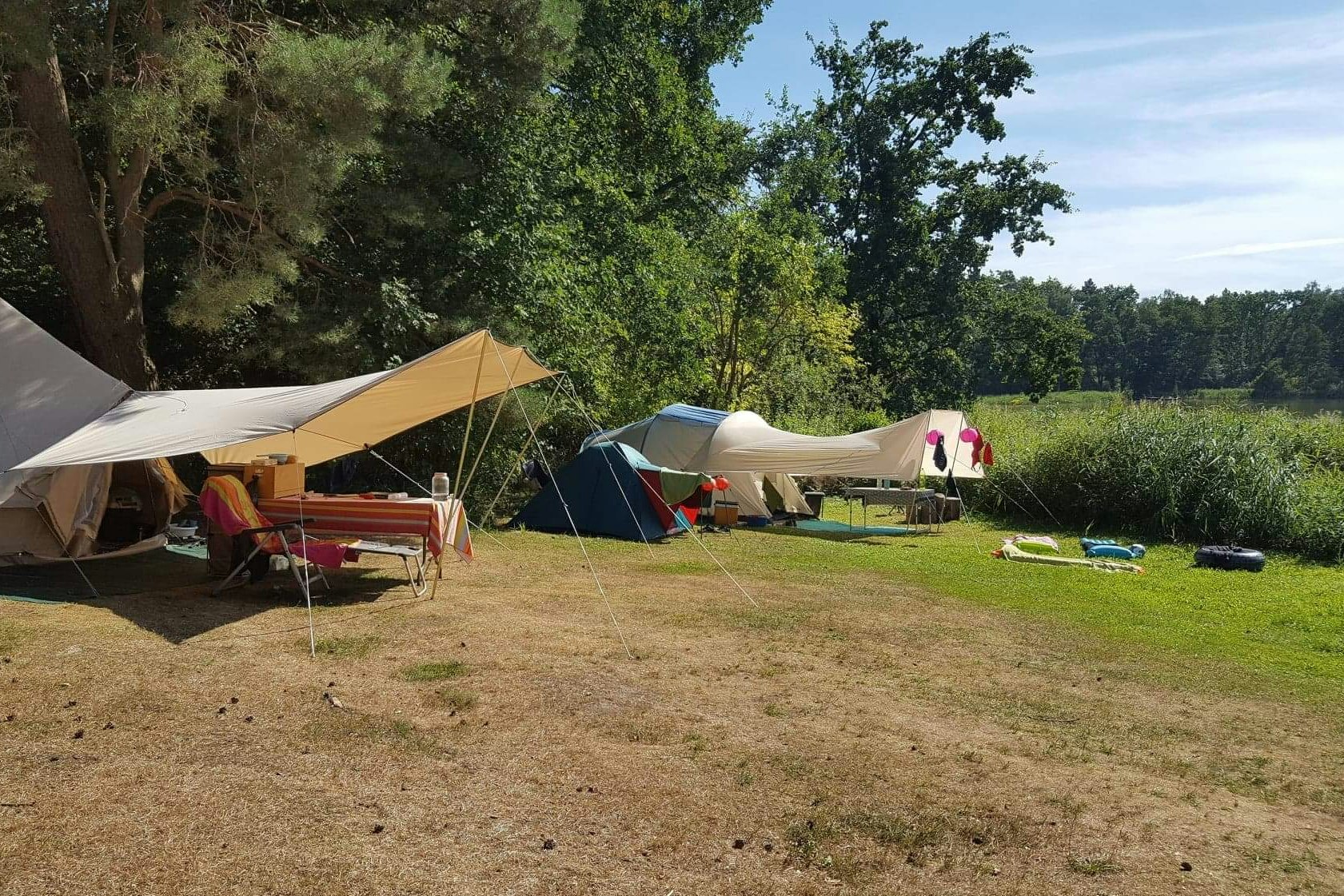 Campingzelte und Sonnensegel auf einem grasigen Stellplatz, mit Stühlen und Liegen im Vordergrund, umgeben von Bäumen.
