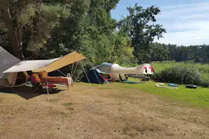 Campingzelte und Sonnensegel auf einem grasigen Stellplatz, mit Stühlen und Liegen im Vordergrund, umgeben von Bäumen.