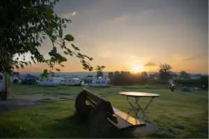 Campingplatz bei Sonnenuntergang: Holztisch und Bank auf der Wiese, im Hintergrund Wohnmobile und Wohnwagen zwischen Bäumen.