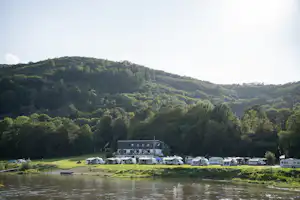 Campingplatz am Fluss mit Wohnmobilen und Zelten, im Hintergrund ein Waldhang und ein Gebäude.