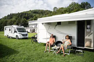 Wohnmobil auf grünem Gras, zwei Personen sitzen am Klapptisch vor dem Eingang, im Hintergrund weitere Fahrzeuge und Bäume.