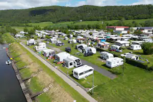 Campingplatz mit vielen Wohnwagen und Wohnmobilen entlang eines Weges, am Flussufer und mit grünen Wiesen zwischen den Stellplätzen.