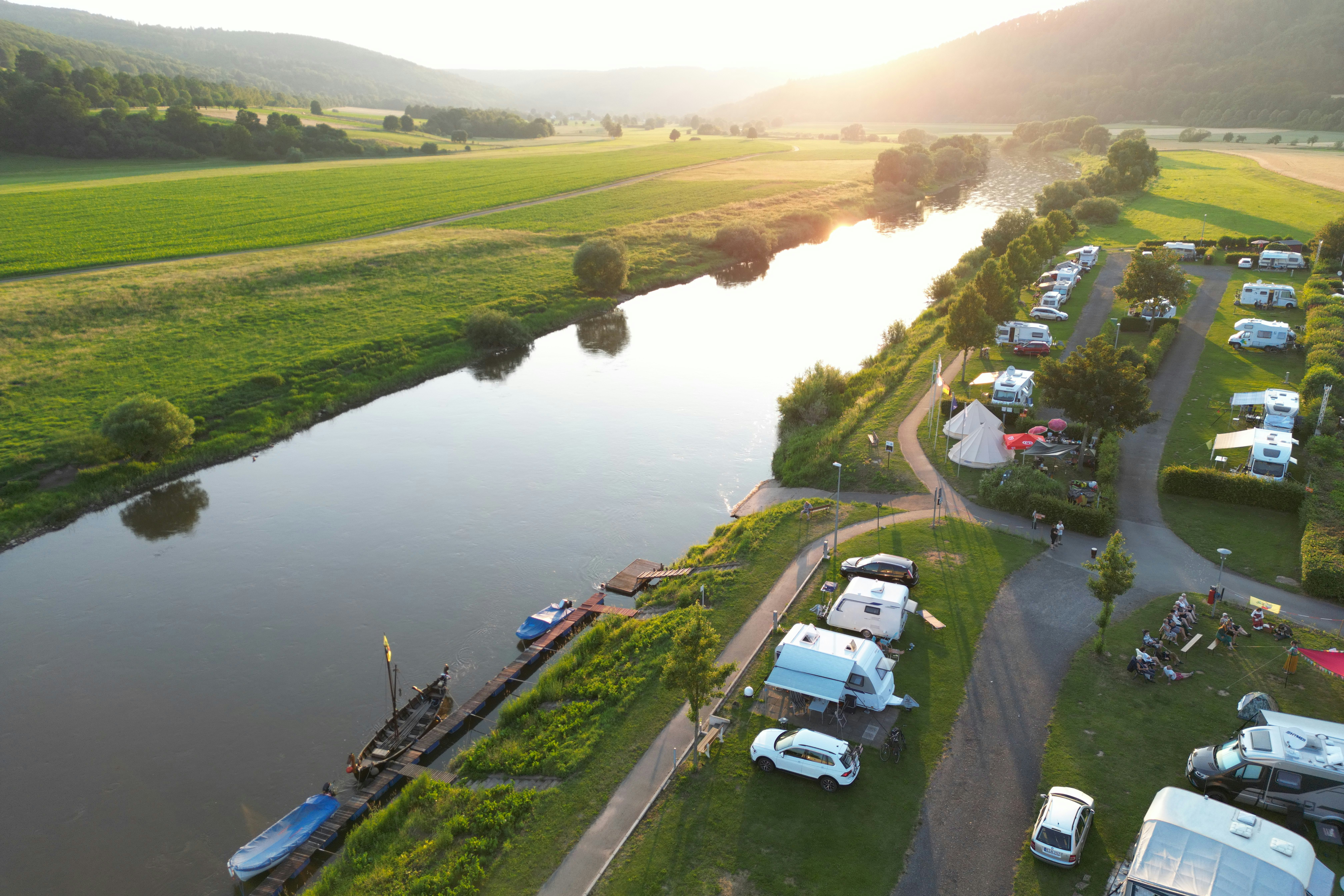 Campingplatz am Fluss mit Wohnmobilen und Stellplätzen entlang des Ufers, umgeben von grünen Feldern und Bäumen.