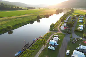 Campingplatz am Fluss mit Wohnmobilen und Stellplätzen entlang des Ufers, umgeben von grünen Feldern und Bäumen.