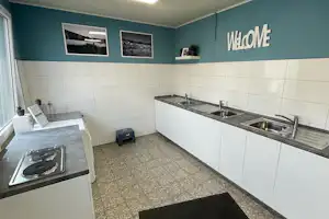 Gemeinschaftsküche mit Spüle und Arbeitsflächen, „WELCOME“-Wandbeschriftung, Fliesenboden und Fensterfront mit Blick auf den Innenraum.