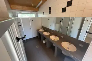 Gemeinschaftsbadezimmer mit mehreren Waschbecken in grauen Schränken, Spiegeln und Fliesenboden, Holzdecke und Fenster im Hintergrund.