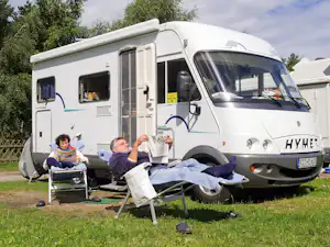 Wohnmobil auf dem Campingplatz, zwei Personen sitzen auf Klappstühlen und lesen vor dem geöffneten Seitenfenster.