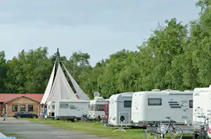Zelt auf dem Campingplatz neben Wohnwagen und Fahrrädern, mit Rasenfläche und Bäumen im Hintergrund.
