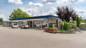 Campinggebäude mit überdachtem Eingang, Stellplätzen für Fahrzeuge und Fahrrädern, davor Betonweg und Blumenbeet.
