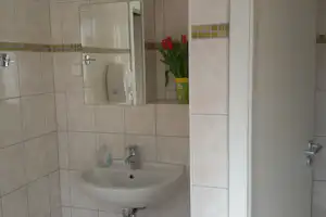 Waschbecken im Sanitärraum mit Fliesenwand, Spiegel und Seifenspendern, daneben eine offene Tür.