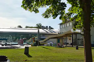 Grüner Rasen mit Baum im Vordergrund, daneben Terrasse und Wohnwagen auf dem Campingplatz, im Hintergrund Gebäude und Zelte.