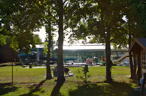 Campingplatz mit Rasen, Sitzgelegenheiten und Bäumen; im Hintergrund ein Gebäude mit Glasfront und Spielbereich.