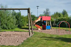 Spielplatz am Campingplatz mit Rutsche, Sandboden und Schaukelgestell, umgeben von Rasen und Bäumen.