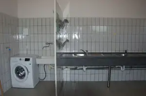 Waschraum mit Waschmaschine, Spülbecken und Armaturen an gefliesten Wänden, dazu Arbeitsplatte und Abflussrohre.